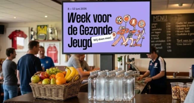 Gezonde kantine met op de voorgrond een mand met vers fruit, flesjes water en bekers met nootjes. Op de achtergrond toont een digitaal scherm de poster van de 'Week voor de Gezonde Jeugd' van 8 tot 12 juni 2026 met de tekst 'Wij doen mee!' en vrolijke illustraties van bewegende kinderen. In de ruimte staan mensen aan de bar.